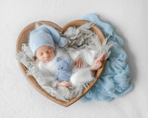 Newborn Baby In White Jumpsuit Sleeps In Wooden Heart-Shaped Bowl During Studio Photoshoot