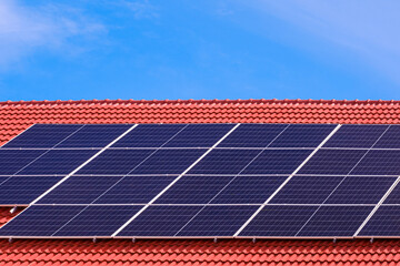 Row of solar panels on top of orange tile roof of modern house against blue sky background,...