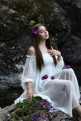 Young woman with long hair in white vintage dress sitting among rocks and stones. Dreaming woman with purple flowers in hair