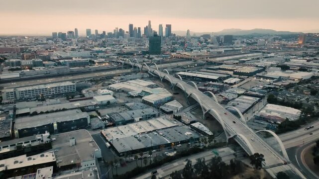 A drone shot of the Ribbon Of Light 6th Street Bridge during sunset in Los Angeles California with cars driving in the foreground of the city's skyline from about 300 feet above the ground