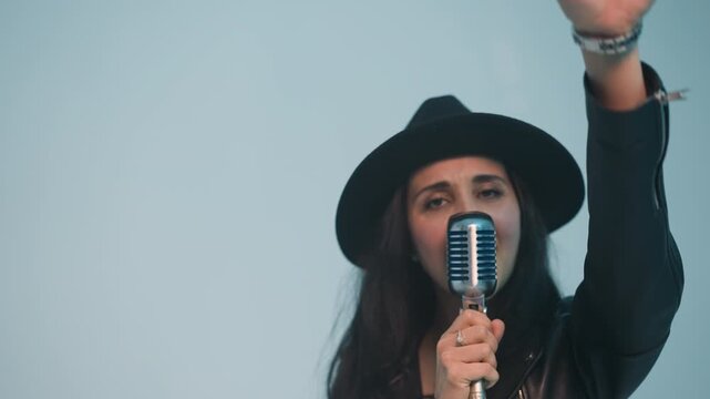 Dynamic front view of a passionate female singer in a black hat and leather jacket, holding a vintage microphone in one hand, moving the other, against a soft white background