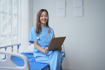 Young doctor is smiling while sitting on a hospital bed and holding a clipboard in her hands