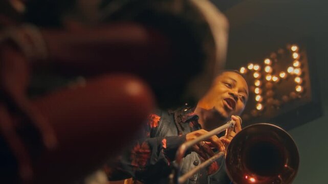 Tilt up shot of Black man dancing, singing and playing trombone with jazz band at concert in bar