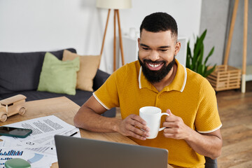 A cheerful man sips coffee while engaged with his laptop at home.