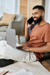 A relaxed man smiles while balancing a phone call and laptop work at home.