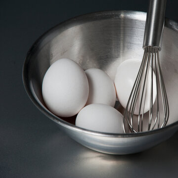 A metal bowl with a balloon whisk and white eggs.