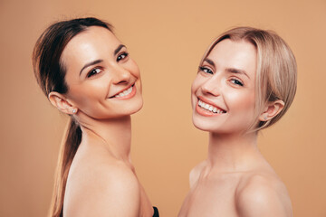 Beauty portrait of two young happy smiling beautiful blonde and brunette women. Models posing naked. Female isolated over beige wall background. Cheerful and happy. Cosmetology concept