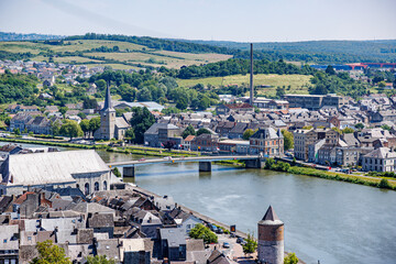 Aerial view of Givet town cityscape, bridge with flags over Meuse river, buildings, houses and churches, field with trees on hill in background, sunny summer day in Ardennes department, France