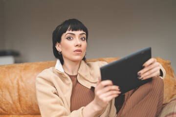 Young Woman Sitting on Couch Engaged With Tablet While Surprised During Indoor Afternoon Activity
