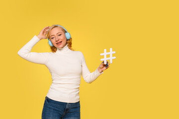 Young Woman in White Sweater Enjoying Music While Holding Hashtag Sign Against Yellow Background