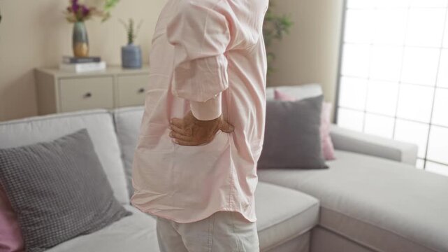 Elderly man experiencing back pain while standing and sitting in a modern living room with a comfortable grey sofa and decorative pillows.
