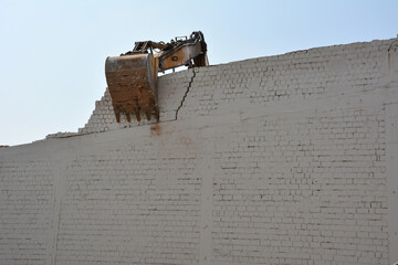 An excavator bucket breaks a brick wall against a blue sky. Only part of the boom and bucket are...