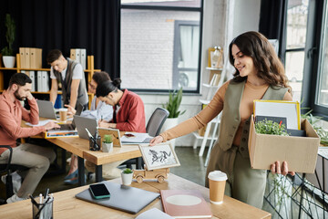 Woman packing belongings during a layoff, colleagues on backdrop.