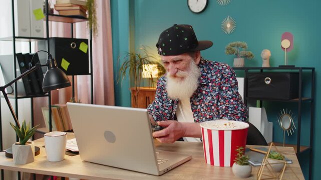 Excited Caucasian senior man celebrating victory in video game on laptop clenching fists, spending leisure time at home office desk. Happy stylish grandpa playing video games holding controller.