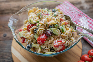 Greek pasta salad with feta cheese, cucumber, tomatoes, olives and onions in a glass bowl isolated on wooden table background