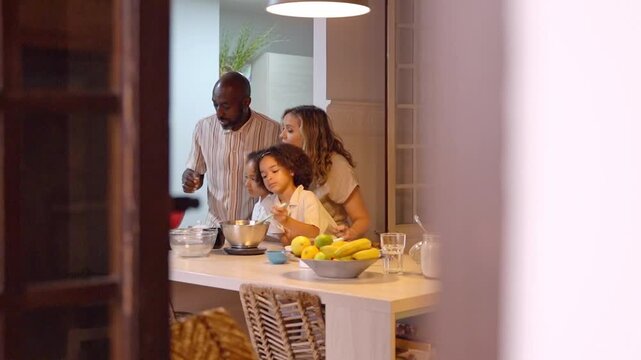 Happy interracial couple teaching little mixed race daughters cooking pancakes and healthy food in modern kitchen evening. Smiling multiracial family preparing with adorable small diverse children