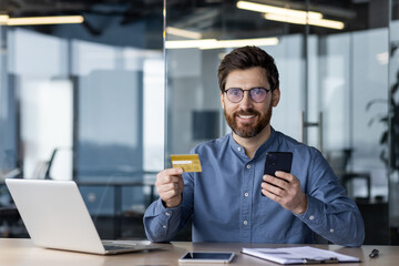 Smiling businessman using smartphone and credit card in modern office with laptop