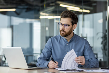 Focused businessman analyzing documents while working on laptop in modern office