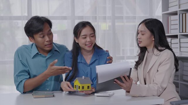 Young couple consults with a real estate agent in a modern office, discussing architectural plans and a house model. Cheerful and engaging conversation