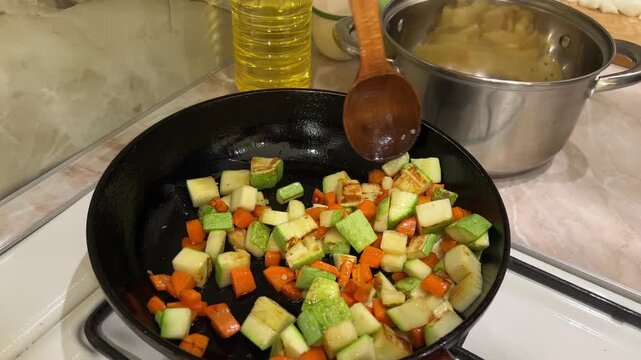 Chef stir frying diced zucchini and carrots in a cast iron skillet on a stovetop with a wooden spoon, preparing a healthy vegetarian meal