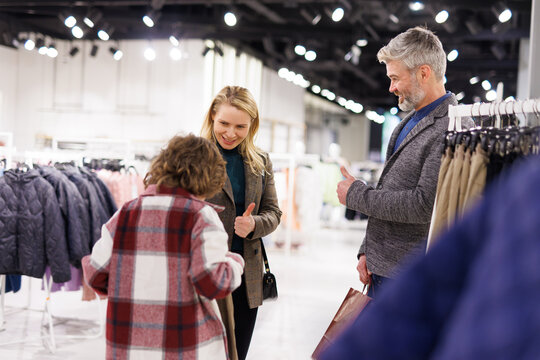 Happy parents showing thumb ups to son choosing new pants in store