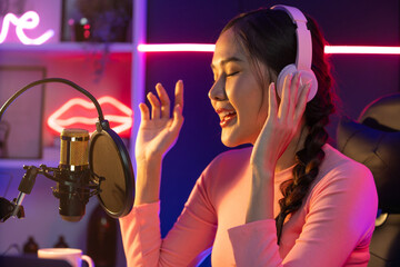 Host channel of young beautiful Asian broadcaster listening the music on social media with listeners, wearing pastel headphones, using microphone for singing at neon light indoor studio. Stratagem.