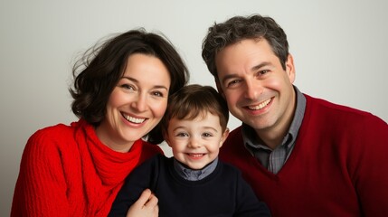 Fototapeta premium Happy family portrait with mother, father, and young child smiling together in matching red and navy outfits against a neutral background.