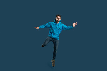 Obraz premium A young man wearing a vibrant blue hoodie is playfully balancing on one leg, smiling widely. The setting features a simple dark background, emphasizing his dynamic pose