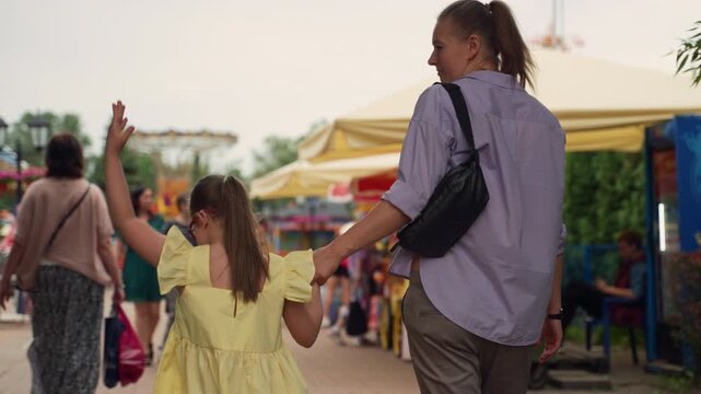 Mother and her young daughter with Down syndrome enter amusement park. Girl in yellow dress points excitedly while holding her mother's hand. Scene highlights parenting child with special needs