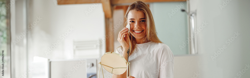 Wall mural Young woman removing makeup with cotton pad in front of mirror standing in bathroom