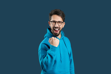A young man wearing a bright blue hoodie expresses excitement with a joyful fist pump. His cheerful...