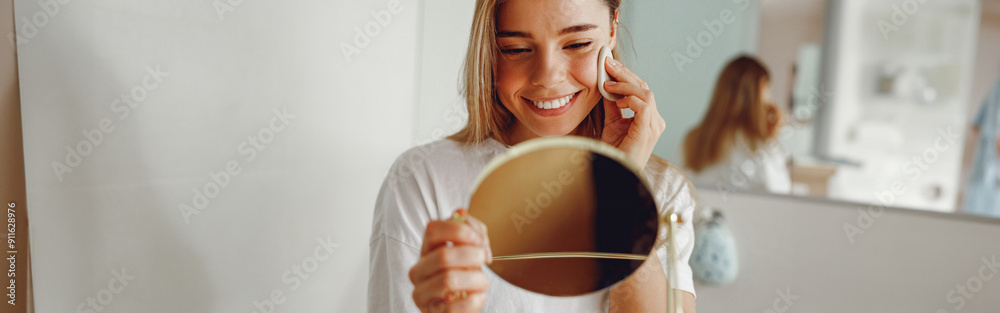 Wall mural Smiling woman removing makeup with cotton pad in front of mirror standing in bathroom