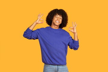 A cheerful man is smiling broadly while making the okay gesture with both hands. The vibrant yellow...