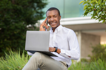 A young African American professional is sitting in a lush green area, actively engaging in a phone conversation while working on a laptop. The individual appears focused and motivated