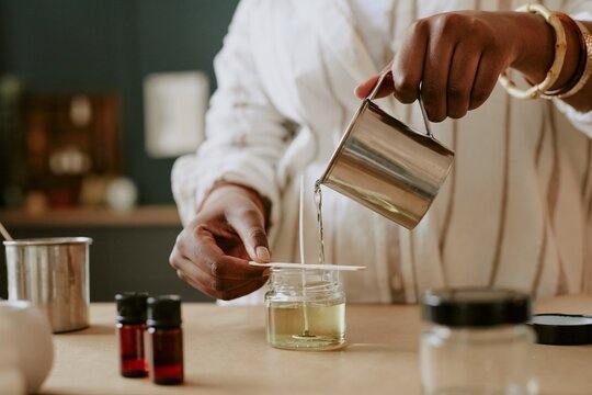 Hands holding a container, pouring wax into a glass jar while crafting handmade candle with essential oils in cozy home setting. Creating a peaceful, aromatic atmosphere