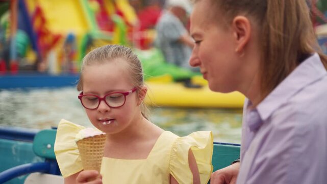 At amusement park, girl with Down syndrome in yellow dress savors ice cream cone as her mother watches. Scene highlights delight of caring for child with special needs