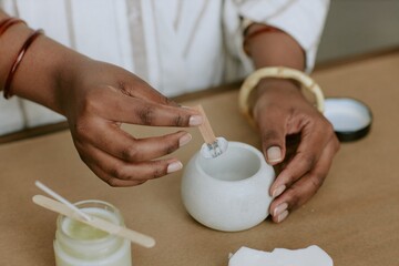 Person stirring mixture in a ceramic container while preparing skincare products at home studio. Person wearing white clothing and bracelets, emphasizing hands and motion