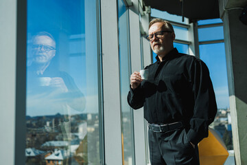 Old business man standing by the window with coffee. He is wearing a black shirt and glasses. A businessman looks at the city from the office window in a skyscraper