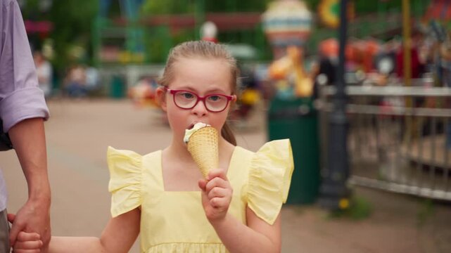Young girl with Down syndrome in yellow dress enjoys eating ice cream cone while sitting with her mother at amusement park. Scene captures simple joys of raising child with special needs.
