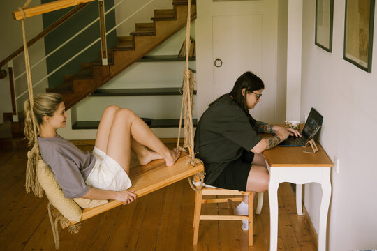 Woman browsing with laptop and partner sitting on swing chair - Powered by Adobe