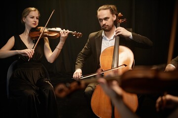 Virtuoso female and male string musicians playing violin and cello on stage with contrast top light against black curtains © Seventyfour