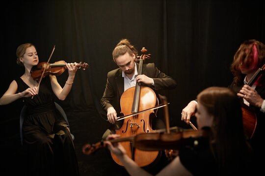 Male classical music artist passionately playing cello with string quartet musicians sitting in circle in studio with contrast top light against black curtains