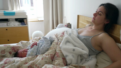Tired mother resting in bed with newborn baby laid in bed trying to regain energy after childbirth,...