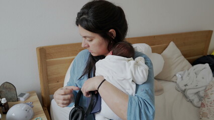 Tired mother sitting down by bedside with newborn baby in arms, lays down to rest after feeling...