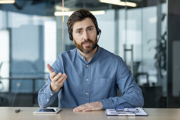 Portrait of a young man in a headset sitting in the office at the table and talking to the camera,...