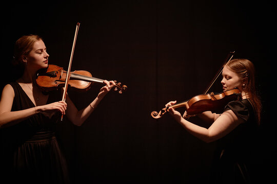 Side view of elegant string duet of female violinists playing classical music in contrast top light on stage with black curtains, copy space