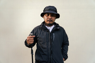 Photo of Asian man wearing all black adventure outfit. Half body portrait of an adult adventurer posing wearing a jacket, boonie hat and trekking poles isolated on a beige background