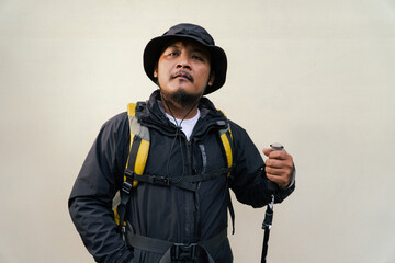 Photo of Asian man wearing adventurer outfit. Half body portrait of an adventurer doing many poses while wearing jacket, hat, backpack and trekking poles isolated on beige background ​