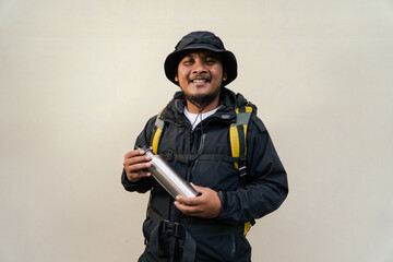 Photo of asian man wearing adventure outfit drinking from stainless steel bottle isolated on beige background. Half body portrait of an adventurer posing drinking from a Tumblr bottle