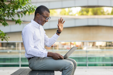 A young African American professional wearing glasses is happily participating in a virtual meeting. He sits on a wooden bench in an outdoor urban area, waving towards the screen of his laptop.